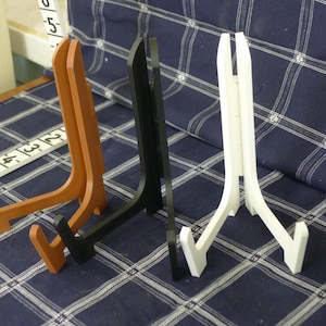 May include: Three display stands in brown, black, and white. Each stand has a three-legged design with a support arm. The stands are made of wood or a similar material and are designed to hold plates or other items.