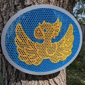May include: A circular wall plaque featuring a silver outer frame and a blue honeycomb mesh background. Centered on the mesh is a yellow, three-dimensional stylised parrot figure with outstretched wings, crafted in a matching honeycomb pattern.