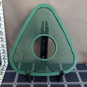 May include: A green, triangular-shaped object with a large circular hole in the center. The object has a honeycomb pattern of small holes throughout. It is supported by a black stand. The object appears to be approximately 6 inches in length.