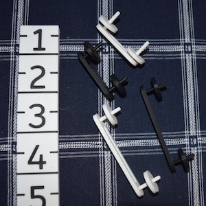 May include: An assortment of white and black plastic components, possibly for a craft or DIY project, are scattered on a blue plaid fabric. A white ruler with black numbers from 1 to 5 is on the left side of the image.