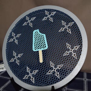May include: A round, blue and silver decorative piece with a honeycomb pattern. It features white star-shaped designs and a light blue popsicle with a bite taken out of it. The popsicle is on a wooden stick.