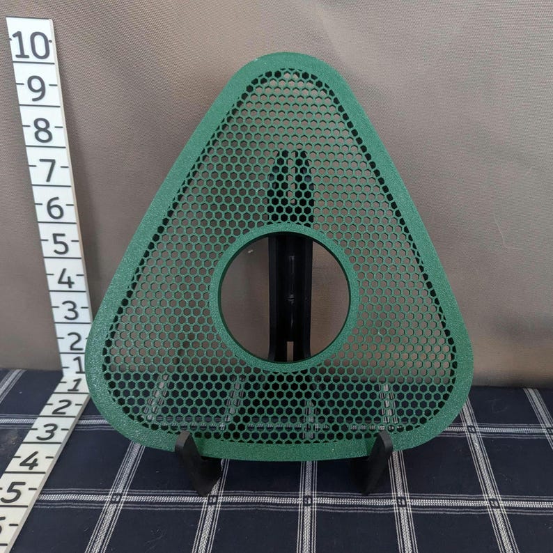 May include: A green, triangular-shaped object with a circular opening and a honeycomb pattern. The object is supported by a black stand. A ruler is placed to the left of the object, showing measurements in inches.