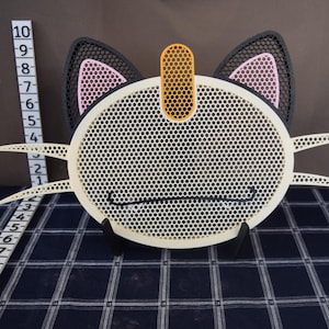 Puede incluir: Un objeto decorativo con forma de gato con una cara blanca perforada, orejas negras con detalles rosas y un acento amarillo. El objeto tiene una altura aproximada de 25 cm.