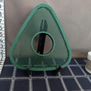 May include: A green, triangular object with a circular hole in the center and a mesh pattern. The object is supported by a black stand. The background is a mix of gray and a blue and white checkered pattern.