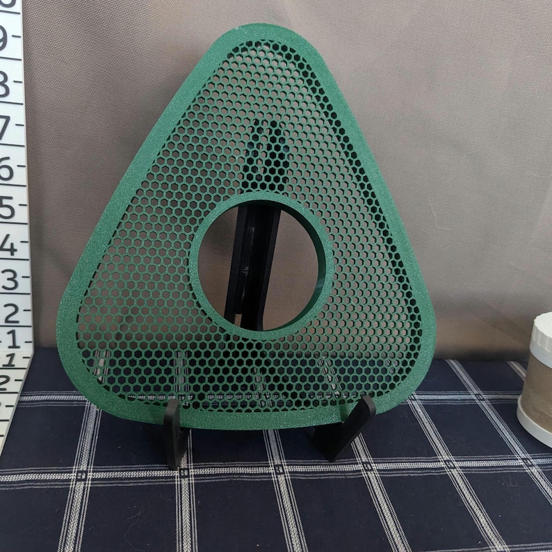 May include: A green, triangular object with a circular hole in the center and a mesh pattern. The object is supported by a black stand. The background is a mix of gray and a blue and white checkered pattern.