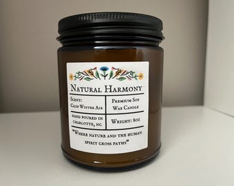 May include: A brown glass candle jar with a black lid. The label reads "NATURAL HARMONY", "SCENT: CRISP WINTER AIR", "PREMIUM SOY WAX CANDLE", "HAND POURED IN CHARLOTTE, NC", "WEIGHT: 8oz", and "WHERE NATURE AND THE HUMAN SPIRIT CROSS PATHS".