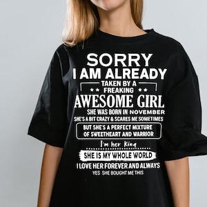 Pode incluir: Camiseta preta com texto branco. O texto diz "SORRY I AM ALREADY TAKEN BY A FREAKING AWESOME GIRL" e outras frases. A camiseta é um presente para quem nasceu em novembro.