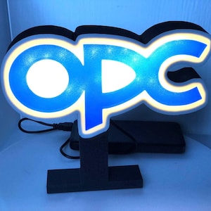 Könnte beinhalten: Ein blaues und weißes LED-Schild mit den Buchstaben "OPC" in einer stilisierten Schriftart. Das Schild steht auf einem schwarzen Ständer und hat ein USB-Kabel.