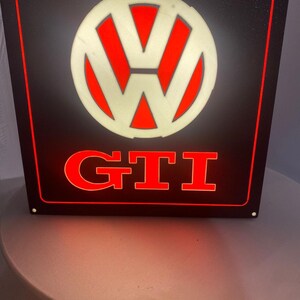 Könnte beinhalten: Ein quadratisches, beleuchtetes schwarzes Schild mit dem weißen Volkswagen-Logo oben und dem roten Schriftzug GTI darunter. Das Schild ist von einem dünnen roten Rand umrahmt und weist kleine Befestigungslöcher in den vier Ecken auf.