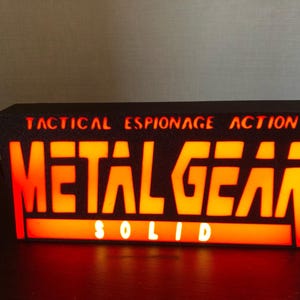 Könnte beinhalten: Ein rechteckiges, schwarzes Leuchtschild mit den Worten "METAL GEAR SOLID" in großen, orangefarbenen, beleuchteten Buchstaben. Oberhalb des Titels steht der Text "TACTICAL ESPIONAGE ACTION" in kleineren, orangefarbenen Buchstaben. Ein Gaming-Sammlerstück.