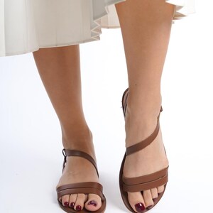 K&ouml;nnte beinhalten: Braune Ledersandalen mit d&uuml;nnen Riemen und Schnallenverschluss. Die Sandalen haben ein minimalistisches Design und werden mit einem wei&szlig;en Faltenrock getragen. Das Bild ist aus einer niedrigen Perspektive aufgenommen, wobei das Schuhwerk hervorgehoben wird.
