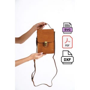 May include: A tan leather crossbody bag with a flap and a gold-coloured clasp. The bag has a long, adjustable strap and is held by a person's hands against a white background. The bag is rectangular and has a small decorative detail.