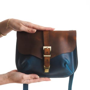 Op de afbeelding: Een tweekleurige leren crossbodytas met een bruine flap en een blauwe body. De tas heeft een messing gespsluiting en een bijpassende blauwe riem. De tas wordt door twee handen omhoog gehouden.