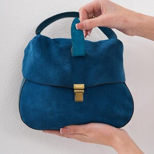 May include: A teal suede handbag with a rounded shape and a top handle. The bag features a flap closure with a gold-colored clasp. The handle is held by a person's hand.