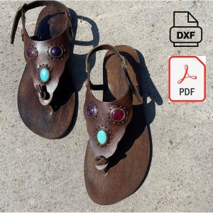 Könnte beinhalten: Ein Paar braune Ledersandalen mit dekorativen Edelsteinen. Jede Sandale hat eine Zehenschlaufe und einen Knöchelriemen. Der obere Teil der Sandalen ist mit lila, roten und türkisfarbenen Steinen verziert. Die Sandalen sind für eine bequeme Passform konzipiert.