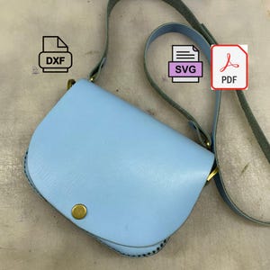May include: A light blue leather crossbody bag with a rounded flap and a long strap. The bag has a gold-colored clasp and a circular button closure. The strap is a darker blue color. The bag is shown with DXF, SVG, and PDF icons.
