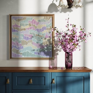 Op de afbeelding: Een abstract kunstwerk in een houten lijst, met een textuur in pastelkleuren, hangt boven een blauwe kast. Een vaas met roze bloemen staat op de kast, naast een wandlamp. Het kunstwerk gebruikt blauw, groen, geel en paars.