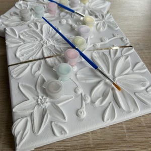 May include: White canvas art with raised floral designs, accompanied by small paint pots in pastel colors and a blue paintbrush. The canvas has a gold stripe. This DIY art kit is perfect for creative projects.