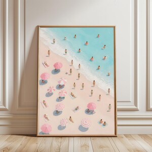 May include: Framed art print of a beach scene with people, pink parasols, and turquoise water. The artwork shows people sunbathing, swimming, and relaxing on the sand. The print is in a wooden frame.