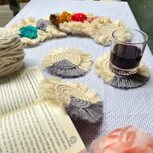 May include: A set of handmade crocheted coasters with a fringe border. The coasters are in various colors, including gray, white, red, orange, yellow, and blue. The coasters are arranged on a white tablecloth with a glass of red wine and a book.