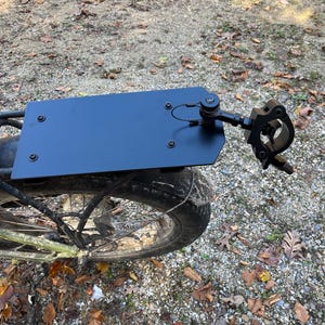 May include: A black metal rack attached to the rear wheel of a bicycle. The rack has a rectangular platform and a mounting bracket. The bicycle tire is black and has a thick tread. The background is a gravel path with fallen leaves.