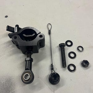 May include: Black metal clamp with adjustable ball joint, a bolt, washers, a nut, and a small cable with loops. The components are arranged on a light gray surface. The clamp has a lever for tightening.