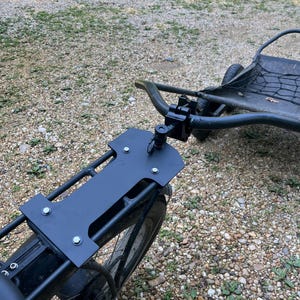 May include: Black metal bicycle rack with a rectangular mounting plate, attached to a bicycle. The rack is bolted to the bicycle frame and has a curved handlebar and a small trailer attached. The bicycle is on a gravel surface.