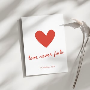 Könnte beinhalten: Eine weiße Karte mit einem roten Herzen und dem Text "love never fails" in roter Kursivschrift. Der Text "1 Korinther 13:8" ist in Schwarz unter dem kursiv geschriebenen Text gedruckt.