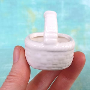 May include: A small, white ceramic basket with a handle. The basket has a woven texture and is held in a person's hand. The background is a light blue and green color.