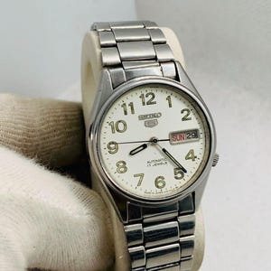 May include: A silver-tone Seiko 5 automatic wristwatch with a cream-colored dial, featuring black Arabic numerals, luminous hands, and a day-date window at the three o'clock position. The watch includes a stainless steel link bracelet.