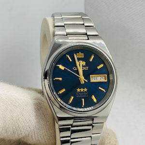 May include: A silver-tone stainless steel wristwatch with a deep blue circular dial, gold-tone hour markers, and gold-tone hands. The watch features a day-date window at the three o'clock position, the Orient logo, and the text Crystal 21 Jewels.