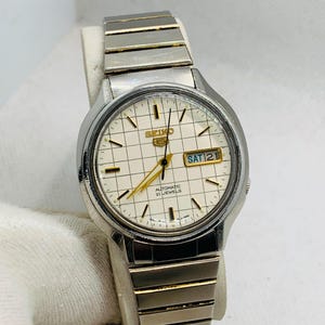 May include: A Seiko 5 automatic wristwatch featuring a cream-colored dial with a grid pattern, gold-tone hands, and hour markers. The watch has a day-date window at the three o'clock position and a two-tone silver and gold stainless steel link band.