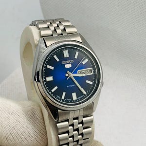 May include: A Seiko 5 automatic wristwatch with a round silver-tone stainless steel case and a matching link bracelet. The watch features a deep blue sunburst dial, silver-tone hour markers, luminous hands, and a day-date window at the 3 o'clock position.