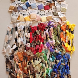May include: A collection of embroidery floss in a variety of colors, including shades of brown, green, blue, red, yellow, and purple. Some floss is still in its original packaging, while other bundles are loose. The image is a top-down view of the floss.