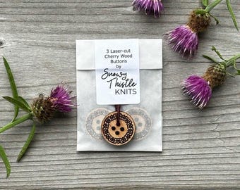 Può includere: Un pacchetto di tre bottoni in legno di ciliegio tagliati al laser di Snowy Thistle Knits. I bottoni sono rotondi con un design decorativo. Il pacchetto è su una superficie di legno, con fiori di cardo viola.