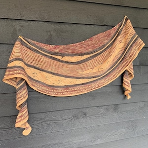 May include: A knitted shawl in peach, grey, and burgundy, featuring horizontal stripes and a detailed openwork border. The shawl is displayed against a dark grey wooden backdrop, highlighting its intricate design and texture.