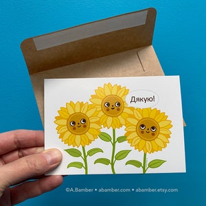 Single cute smiling Ukrainian Sunflowers Thank You Card with kraft paper envelope - held in hand in front of a blue wall. Illustration by illustrator Adrianna Bamber.