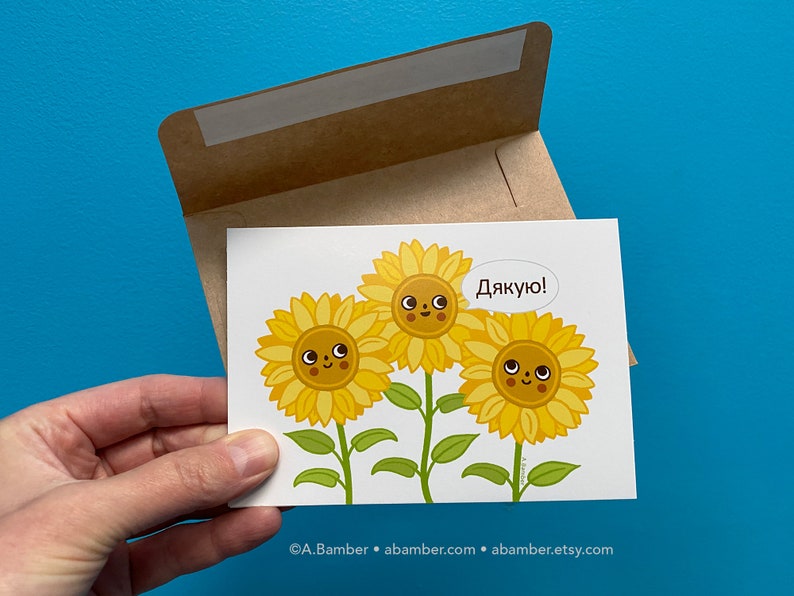 Single cute smiling Ukrainian Sunflowers Thank You Card with kraft paper envelope - held in hand in front of a blue wall. Illustration by illustrator Adrianna Bamber.