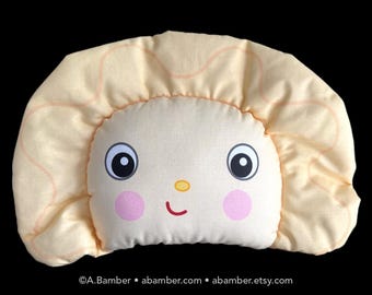 May include: A yellow and peach-colored plush pillow shaped like a smiling face with large eyes and pink cheeks. The pillow has a ruffled edge around the outside.