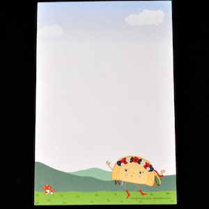 May include: A notepad featuring a whimsical illustration of a smiling taco character wearing a floral crown and red boots, set against a backdrop of a green field, rolling hills, and a blue sky with clouds. The text "abamber.etsy.com" is at the bottom.