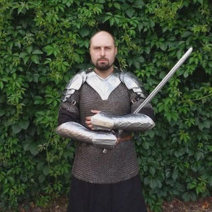 Könnte beinhalten: Eine Person trägt mittelalterliche Rüstung, darunter ein Kettenhemd, Schulter- und Armschützer aus Metall und einen schwarzen Rock. Die Person hält ein langes Silberschwert. Der Hintergrund ist eine Wand aus grünen Blättern.