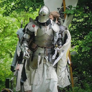 Puede incluir: Un grupo de personas con armadura medieval, incluyendo cascos, petos y protectores de piernas, caminan por una zona boscosa. Una persona lleva una bandera con un emblema negro. La armadura es plateada y la ropa es blanca.