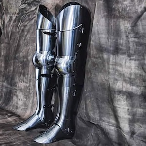 May include: Pair of polished steel leg armor, reflecting the light. The armor features articulated knee joints and is designed to protect the legs. The metal has a shiny, reflective surface, and the armor is designed for historical reenactment or display.