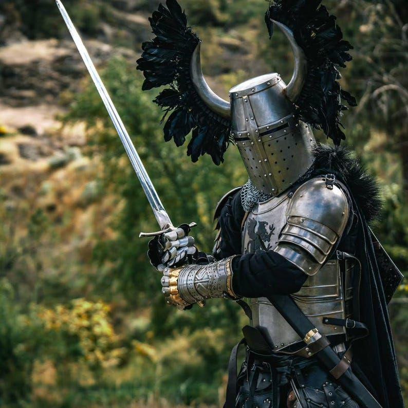 May include: A knight in full armor, holding a long silver sword. The helmet features a horned design with black feathers. The armor is silver with black accents, and a black cape is draped over the shoulders. The background is a blurred forest.