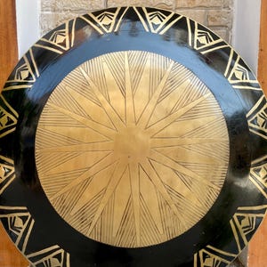 Op de afbeelding: Een rond, zwart en gouden schild met een stervormig ontwerp in het midden. Het schild heeft een zwarte buitenrand met een gouden geometrisch patroon.