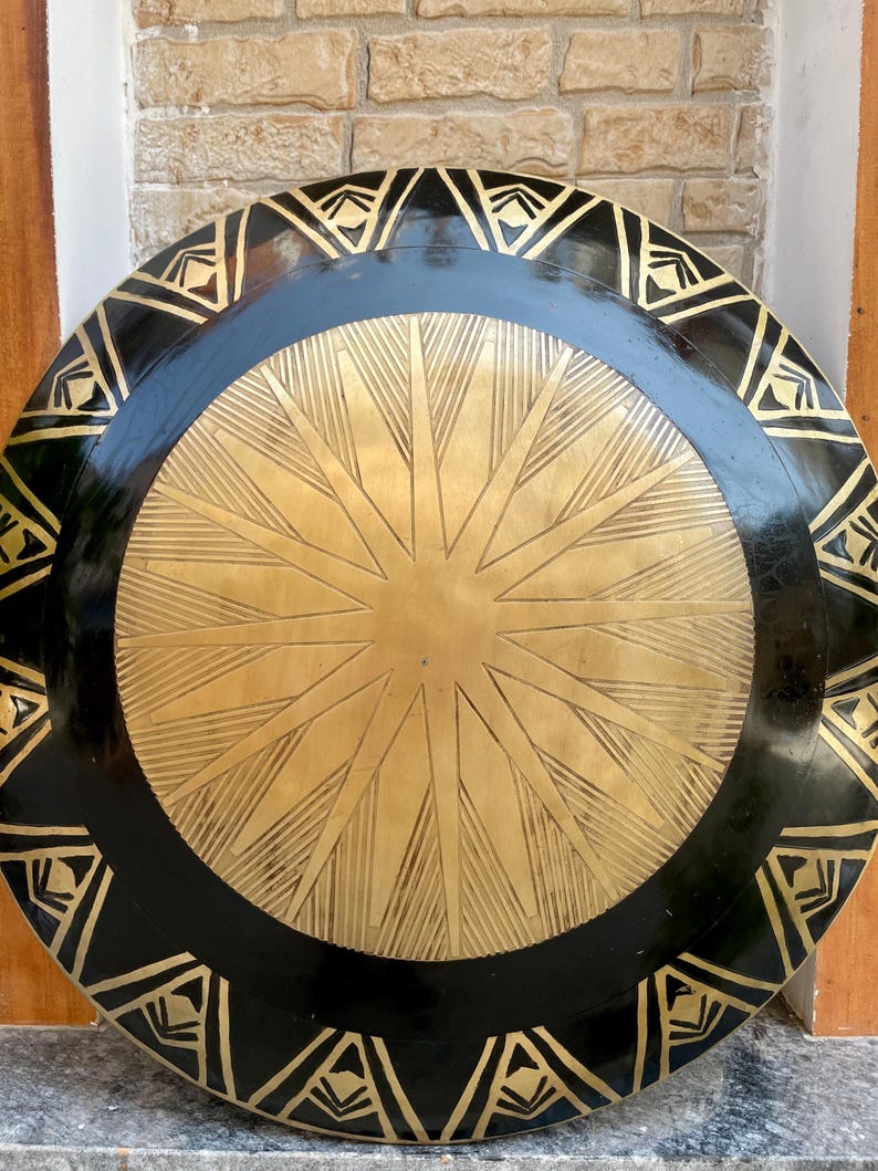 Op de afbeelding: Een rond, zwart en gouden schild met een stervormig ontwerp in het midden. Het schild heeft een zwarte buitenrand met een gouden geometrisch patroon.