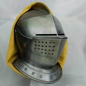 Puede incluir: Un yelmo de caballero de acero pulido con visera y orificios de ventilación. El yelmo está parcialmente cubierto por una capucha amarilla. El yelmo tiene un collar decorativo con remaches.
