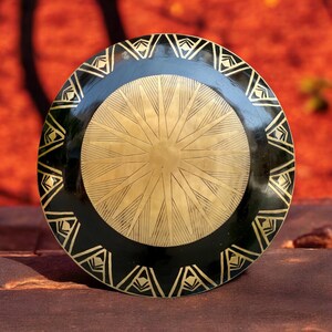 Op de afbeelding: Een ronde, zwart-gouden decoratieve plaat met een geometrisch ontwerp. De plaat heeft een verhoogd midden met een stervormig patroon.