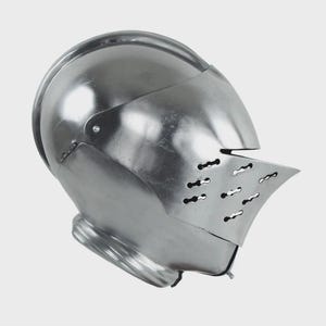 Può includere: Un elmo medievale lucido, color argento, con visiera. L'elmo presenta una parte superiore arrotondata e una visiera incernierata con diverse piccole fessure di ventilazione orizzontali. La superficie metallica riflette la luce, evidenziando il suo design liscio e curvo.