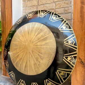 Op de afbeelding: Een groot, rond, zwart en gouden schild met een decoratief ontwerp. Het schild heeft een centraal stervormig patroon en een rand van driehoeken.